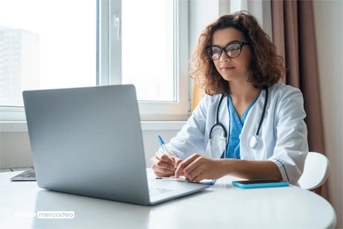 Médica con bata blanca y estetoscopio trabajando en una computadora portátil en su consultorio